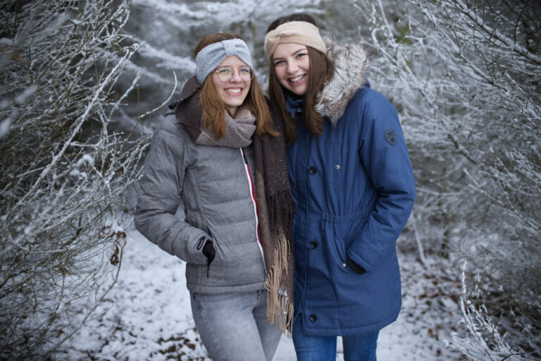 Zwei Freundinnen halten sich im Arm und lächeln in die Kamera. Sie stehen in einem wunderschönen Winterwald