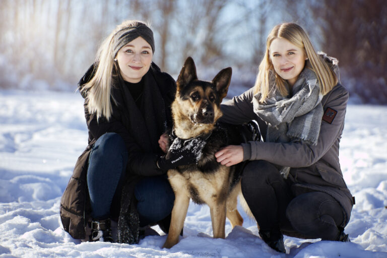 Zwei Mädchen sitzen links und rechts neben ihrem Schäferhund und halten ihn im Arm.